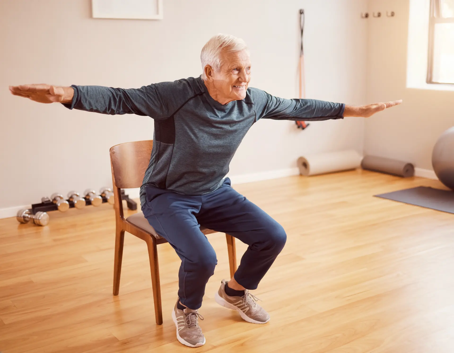 Older man performing a supported squat beside a chair.
